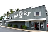 A gray wooden building with a large sign reading 'BAKERY' and established in 1985 is prominently displayed. The front of the building has white-framed windows and a glass door with a person entering. There are palm trees in the background and some greenery near the entrance.