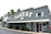 A gray wooden building with a large sign reading 'BAKERY' and established in 1985 is prominently displayed. The front of the building has white-framed windows and a glass door with a person entering. There are palm trees in the background and some greenery near the entrance.