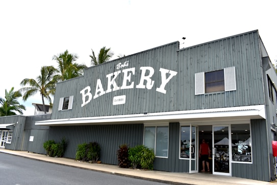 A gray wooden building with a large sign reading 'BAKERY' and established in 1985 is prominently displayed. The front of the building has white-framed windows and a glass door with a person entering. There are palm trees in the background and some greenery near the entrance.
