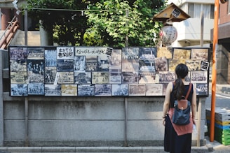 A woman stands in front of a display board covered with a collection of vintage black and white photographs. The board is situated outdoors against a stone wall and is surrounded by lush greenery and urban buildings. The woman has a backpack and is looking at the photographs, which depict various group and individual scenes.