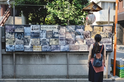 A woman stands in front of a display board covered with a collection of vintage black and white photographs. The board is situated outdoors against a stone wall and is surrounded by lush greenery and urban buildings. The woman has a backpack and is looking at the photographs, which depict various group and individual scenes.