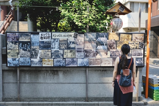 A woman stands in front of a display board covered with a collection of vintage black and white photographs. The board is situated outdoors against a stone wall and is surrounded by lush greenery and urban buildings. The woman has a backpack and is looking at the photographs, which depict various group and individual scenes.
