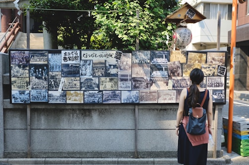 A woman stands in front of a display board covered with a collection of vintage black and white photographs. The board is situated outdoors against a stone wall and is surrounded by lush greenery and urban buildings. The woman has a backpack and is looking at the photographs, which depict various group and individual scenes.