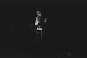 A person in a dark setting, wearing a jacket and gloves, appears to be in a contemplative or prayerful pose with hands together.
