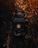 A vintage camera with a large lens is centered against a backdrop of dried, textured grass. The lighting creates a warm, nostalgic ambiance, illuminating the camera's details and casting shadows on the straw-like material.