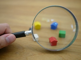 A hand holds a magnifying glass focusing on small, colorful toy houses scattered on a wooden surface. The toy houses are in different colors, including red, blue, yellow, green, and white, and the magnifying glass enlarges their appearance.