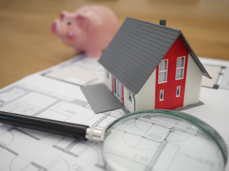 3D home model sitting on top of an architect blueprint with a magnifying glass and piggy bank