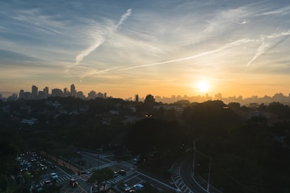 A cityscape at sunrise with the sun rising behind a skyline of tall buildings. The sky is filled with wispy clouds illuminated by the sunrise, creating a gradient of colors. Below, a network of roads and junctions is visible, with trees and traffic starting to build up.