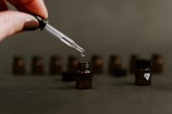 A dropper held by a hand above a small brown glass bottle, with other similar bottles blurred in the background. The focus is on the dropper releasing a droplet into one bottle, while another bottle has a white heart symbol on its cap.