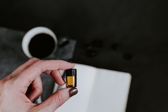 A hand holding a small amber-colored vial in focus, with a notebook and a cup of black coffee in the blurred background.