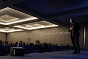 A man is standing on stage in a dimly lit conference room, gesturing as he is speaking to an audience. The room is filled with rows of seated people, and there is a large logo and the words 'Product School' projected on the wall. Overhead lights create a warm ambiance, and there is a speaker placed on the floor of the stage.