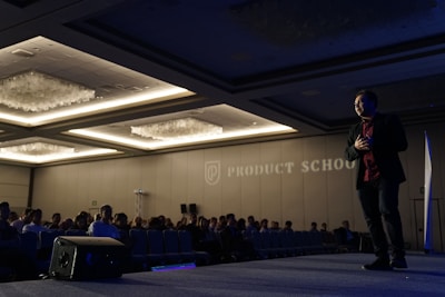 A man is standing on stage in a dimly lit conference room, gesturing as he is speaking to an audience. The room is filled with rows of seated people, and there is a large logo and the words 'Product School' projected on the wall. Overhead lights create a warm ambiance, and there is a speaker placed on the floor of the stage.