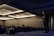 A man is standing on stage in a dimly lit conference room, gesturing as he is speaking to an audience. The room is filled with rows of seated people, and there is a large logo and the words 'Product School' projected on the wall. Overhead lights create a warm ambiance, and there is a speaker placed on the floor of the stage.