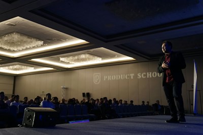 A man is standing on stage in a dimly lit conference room, gesturing as he is speaking to an audience. The room is filled with rows of seated people, and there is a large logo and the words 'Product School' projected on the wall. Overhead lights create a warm ambiance, and there is a speaker placed on the floor of the stage.