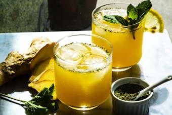 Two glasses of a yellow, refreshing beverage with ice, garnished with mint leaves and a slice of lemon, are set on a table. Beside them, ginger root and fresh mint are displayed, along with a small bowl filled with a green herb and a spoon.