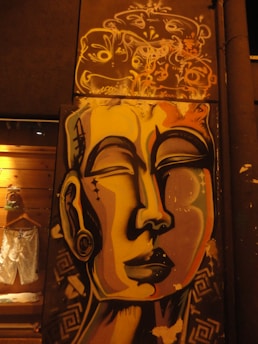 A vibrant mural of a stylized face, featuring bold lines and a serene expression, painted on an urban wall. Above the face is intricate abstract graffiti with swirling patterns and faces. The surrounding environment includes a wooden wall with some clothing hanging on the left side.