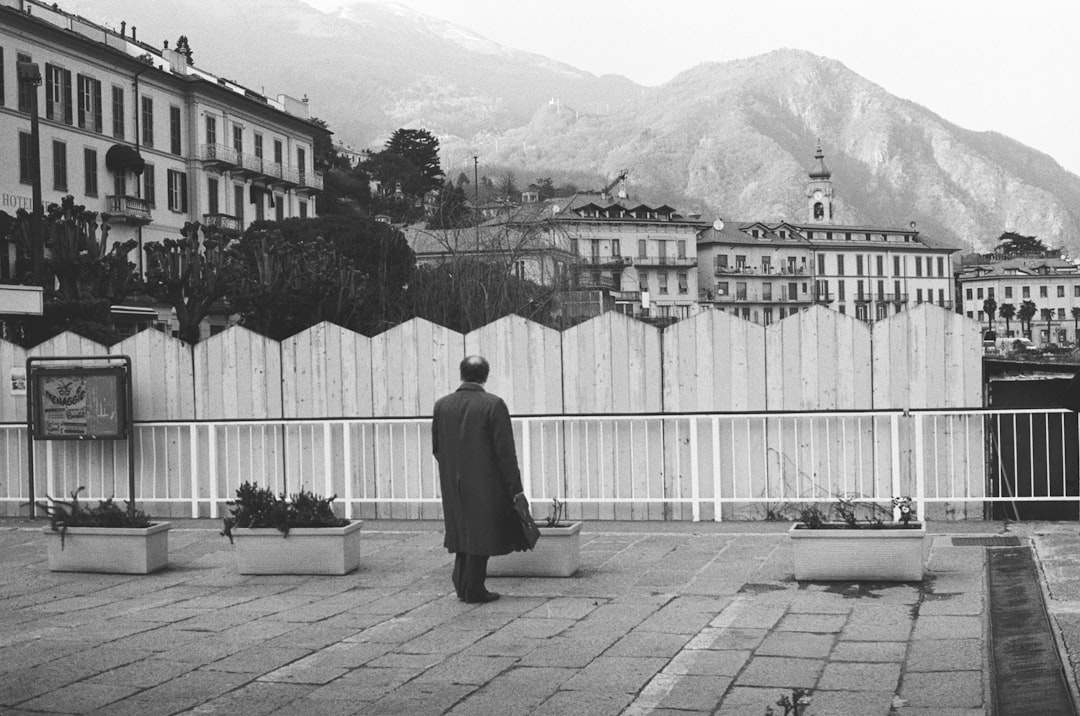 grayscale photo of man in trench coat standing in front of wooden fence