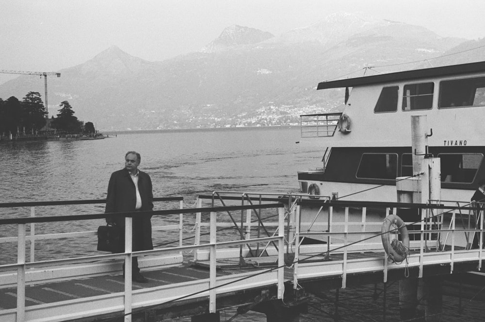 greyscale photo of man standing on dock near boat