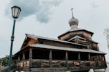A wooden building with a traditional Russian architectural style, featuring a prominent onion dome on top. The structure is aged, with visible wood grains and textures on the exterior. A tall, vintage-style street lamp is positioned in the foreground on the left side, set against a slightly overcast sky.
