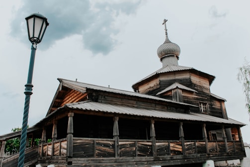 A wooden building with a traditional Russian architectural style, featuring a prominent onion dome on top. The structure is aged, with visible wood grains and textures on the exterior. A tall, vintage-style street lamp is positioned in the foreground on the left side, set against a slightly overcast sky.