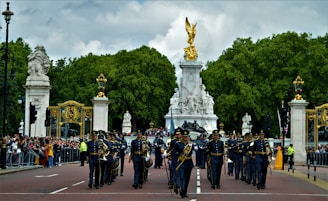 A military marching band is parading down a wide road lined with spectators behind barriers. Large ornate gates and a prominent statue featuring a golden figure form the backdrop against a dense covering of trees and a cloudy sky.