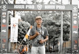 A person stands smiling in a casual pose beneath an archway with the words 'KRUI PRO' displayed prominently. They are holding a camera and wearing a cap, set against a background of trees and motorcycles.