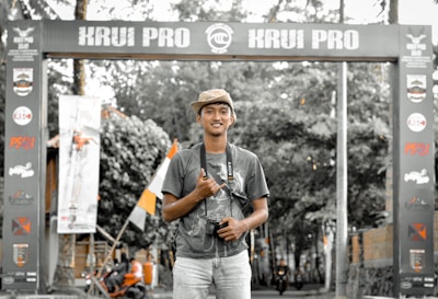 A person stands smiling in a casual pose beneath an archway with the words 'KRUI PRO' displayed prominently. They are holding a camera and wearing a cap, set against a background of trees and motorcycles.
