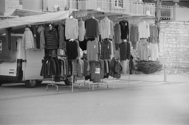 A street market setup features a variety of clothing items hanging on racks and hooks. There is a mix of sweaters, jackets, and other garments displayed on a mobile stall attached to a van. The scene appears to be outdoors with a stone wall and a paved surface visible in the background.
