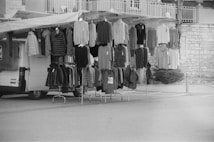 A street market setup features a variety of clothing items hanging on racks and hooks. There is a mix of sweaters, jackets, and other garments displayed on a mobile stall attached to a van. The scene appears to be outdoors with a stone wall and a paved surface visible in the background.