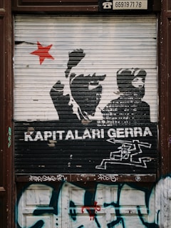 A graffiti mural on a metal shutter features silhouettes of protesters with raised fists and a prominent red star to the left. The words 'KAPITALARI GERRA' are painted in bold white text beneath the figures. Below the text is a stylized image of what appears to be machinery or a weapon. The bottom part of the shutter and surrounding area are covered in additional graffiti tags.
