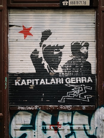 A graffiti mural on a metal shutter features silhouettes of protesters with raised fists and a prominent red star to the left. The words 'KAPITALARI GERRA' are painted in bold white text beneath the figures. Below the text is a stylized image of what appears to be machinery or a weapon. The bottom part of the shutter and surrounding area are covered in additional graffiti tags.