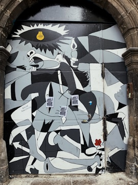 A mural inspired by Picasso's Guernica features abstract forms and distorted figures in a monochromatic palette. The artwork is painted on a large door set within an arched stone frame. Notable elements include a horse, a bull, a light bulb resembling an eye, and several human-like figures intertwined in dynamic poses. Some printed materials are affixed to the middle of the mural.