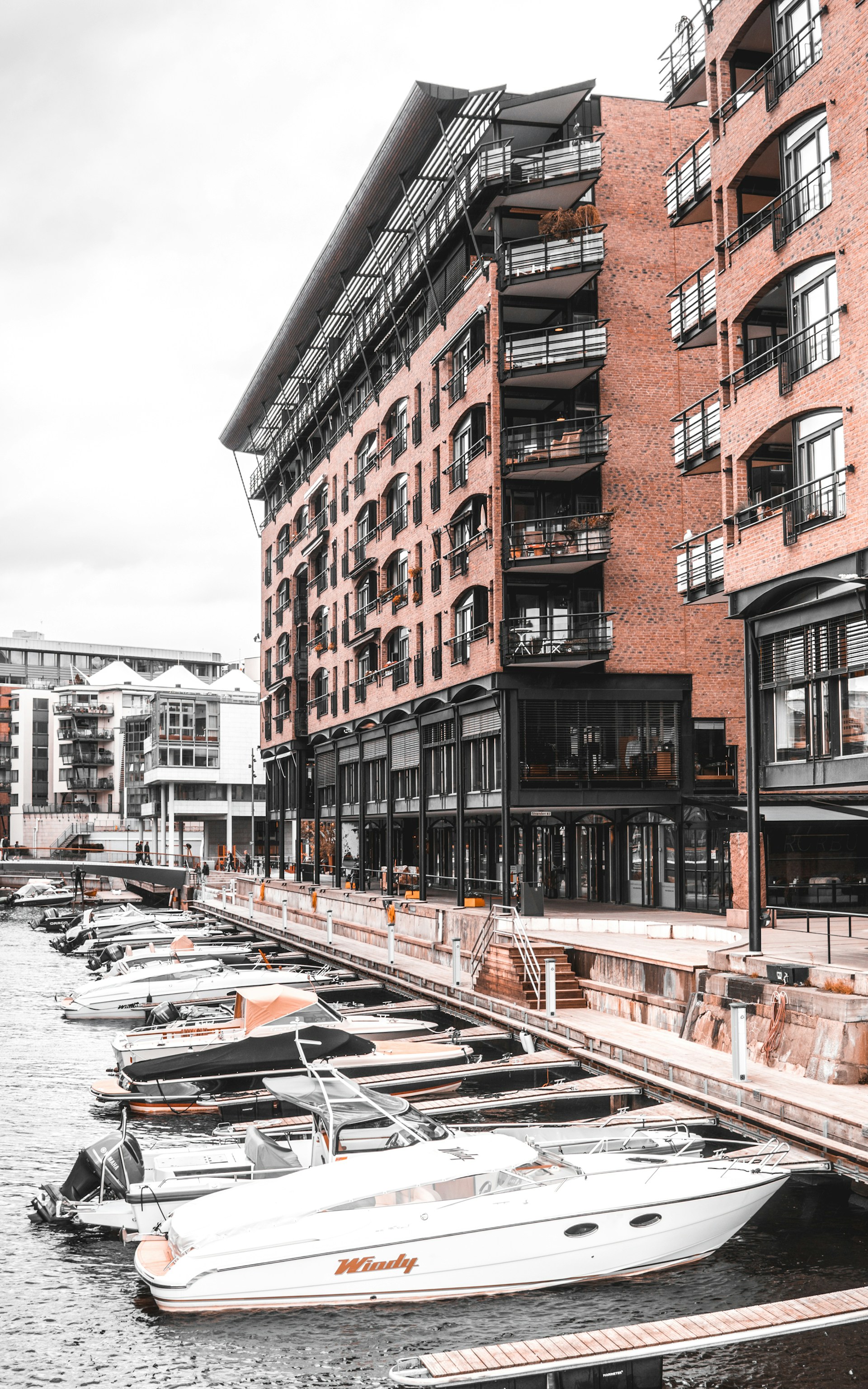 A waterfront scene with modern brick buildings featuring multiple balconies and large windows. There are several moored motorboats along a dockside promenade, with a sleek white boat in the foreground. The setting appears to be urban, with other modern buildings visible in the background.