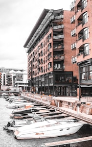 A waterfront scene with modern brick buildings featuring multiple balconies and large windows. There are several moored motorboats along a dockside promenade, with a sleek white boat in the foreground. The setting appears to be urban, with other modern buildings visible in the background.