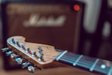 A close-up view of an electric guitar’s headstock with tuning pegs in focus, featuring wooden texture and metallic finishes. In the background, a blurred guitar amplifier is visible, suggesting a musical environment.