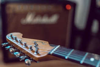 A close-up view of an electric guitar’s headstock with tuning pegs in focus, featuring wooden texture and metallic finishes. In the background, a blurred guitar amplifier is visible, suggesting a musical environment.