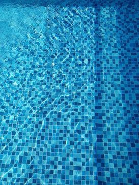 A close-up view of a swimming pool with clear blue water and a tiled bottom. The sunlight creates intricate patterns and reflections on the water surface, enhancing the vibrant blue tones of the tiles.