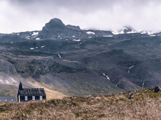 A small black church with a pointed roof stands alone in a vast, open landscape. The foreground is filled with grasslands, and the backdrop features majestic, rugged mountains partially covered in snow. The sky is overcast, lending a dramatic and moody atmosphere to the scene.