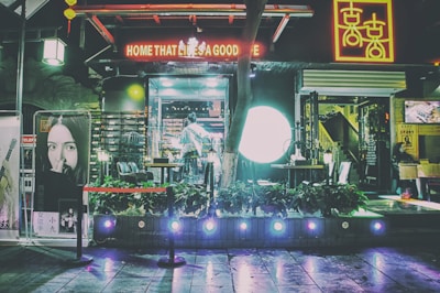 A dimly lit street scene in front of an entertainment venue or cafe, with neon signs in red and yellow. The entrance features glass doors and is surrounded by vibrant lights and decorations. A poster of a woman is displayed outside next to artificial plants and seating arrangements.