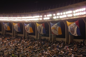 A crowded stadium with an illuminated upper section featuring large, colorful flags that hang between the bright lights. The seats are filled with spectators, hinting at an ongoing or upcoming event.