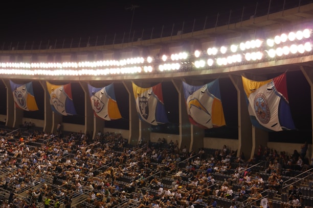 A crowded stadium with an illuminated upper section featuring large, colorful flags that hang between the bright lights. The seats are filled with spectators, hinting at an ongoing or upcoming event.