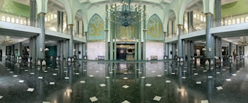 An expansive, ornate interior of a mosque featuring large Arabic calligraphy on the walls, numerous pillars with intricate designs, and a vast marble floor. The ceiling is adorned with a large, elaborate chandelier. Natural light filters in through multiple arched windows creating a serene and sacred atmosphere. The symmetrical architecture and decoration emphasize a sense of grandeur and spirituality.
