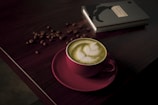A cup of matcha latte with delicate latte art sits on a dark wooden table. The pink cup contrasts with the green frothy beverage. Coffee beans are scattered around near a closed black notebook, adding to the cozy ambiance.