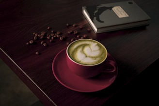 A cup of matcha latte with delicate latte art sits on a dark wooden table. The pink cup contrasts with the green frothy beverage. Coffee beans are scattered around near a closed black notebook, adding to the cozy ambiance.