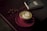 A cup of matcha latte with delicate latte art sits on a dark wooden table. The pink cup contrasts with the green frothy beverage. Coffee beans are scattered around near a closed black notebook, adding to the cozy ambiance.