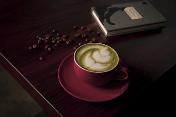 A cup of matcha latte with delicate latte art sits on a dark wooden table. The pink cup contrasts with the green frothy beverage. Coffee beans are scattered around near a closed black notebook, adding to the cozy ambiance.