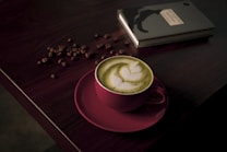 A cup of matcha latte with delicate latte art sits on a dark wooden table. The pink cup contrasts with the green frothy beverage. Coffee beans are scattered around near a closed black notebook, adding to the cozy ambiance.
