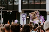 A crowd is gathered in a protest or demonstration. People are holding signs, one featuring the female gender symbol, and another with the word 'Hässig.' The atmosphere suggests activism, with participants wearing casual clothing. In the background, there is a store with mannequins on display.