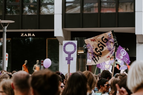 A crowd is gathered in a protest or demonstration. People are holding signs, one featuring the female gender symbol, and another with the word 'Hässig.' The atmosphere suggests activism, with participants wearing casual clothing. In the background, there is a store with mannequins on display.