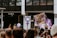 A crowd is gathered in a protest or demonstration. People are holding signs, one featuring the female gender symbol, and another with the word 'Hässig.' The atmosphere suggests activism, with participants wearing casual clothing. In the background, there is a store with mannequins on display.